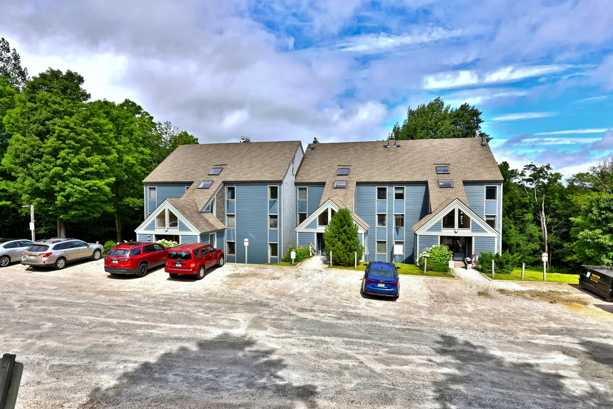 A wide-angle view of two multi-story buildings surrounded by greenery and parked vehicles, suggesting a safe, community-oriented environment. The setting is tranquil, with families visible enjoying outdoor relaxation, morning coffees on the porch, and children playing on the lawn.