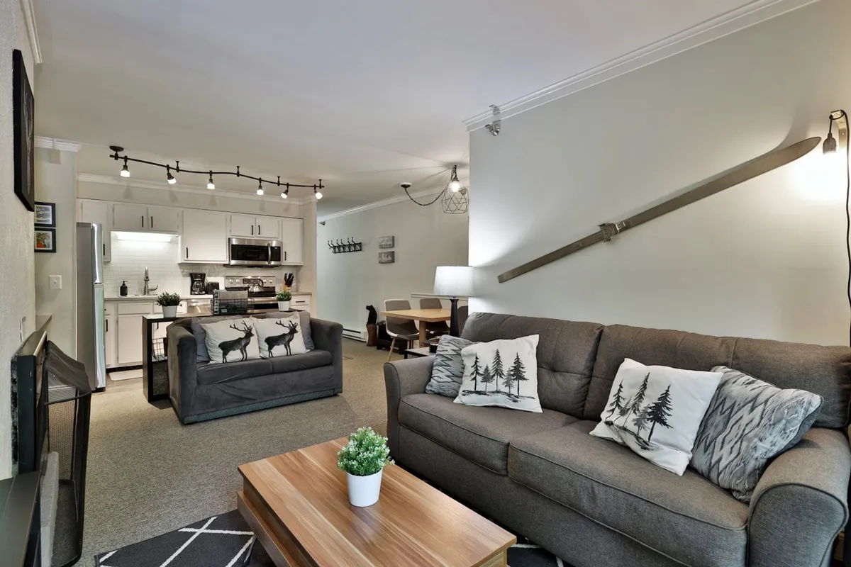A modern, inviting open space with a sofa decorated with forest-themed cushions, a wooden coffee table, an open kitchen area, and a dining space. The layout encourages gathering, sharing stories, and communal meals.