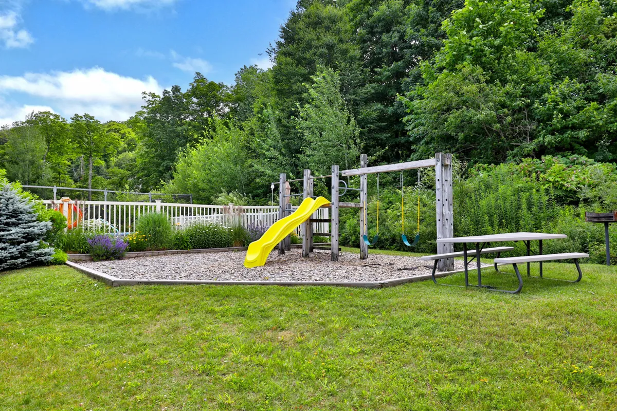 This image features a cheerful playground area with bright yellow slides, swings, picnic tables, and lush greenery. Children are depicted enjoying the equipment, while families gather for picnics nearby. The scene radiates joy, activity, and togetherness in an outdoor setting.