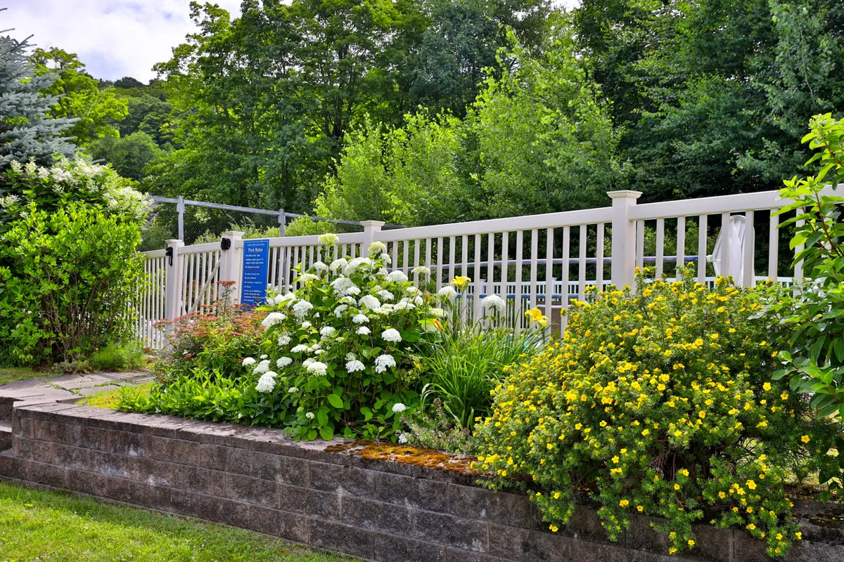 A serene exterior with lush greenery, flowering shrubs, and a white fence. The landscape suggests seasonal changes, evoking tranquility but also the possibility of picturesque winter scenes when snow blankets the area.