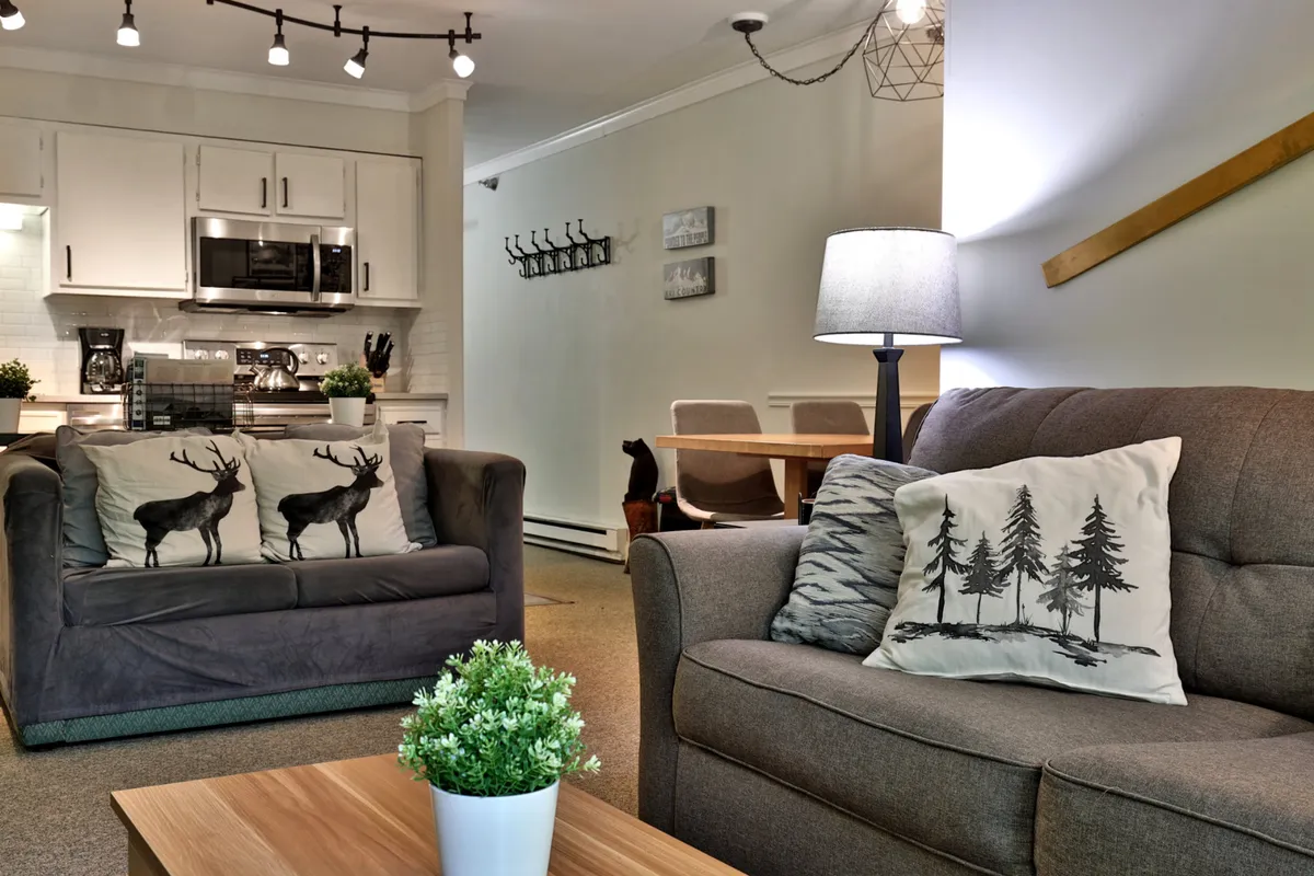 A wide view of an open-plan living area with plush sofas adorned with decorative pillows, a wooden coffee table, a visible kitchen area in the background, a dining table, and hanging light fixtures. The space feels welcoming and comfortable with ample seating for gatherings.