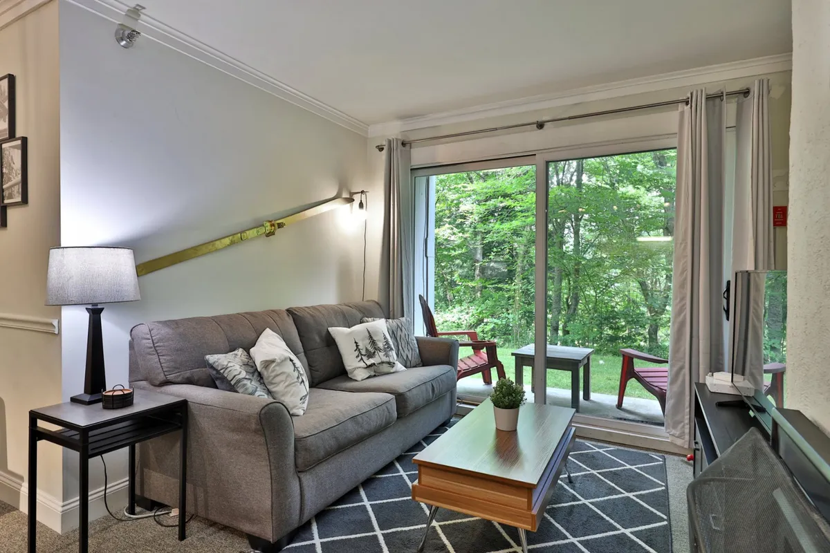 A bright living area with a comfortable sofa, coffee table, and large windows offering views of outdoor seating. The arrangement suggests a flexible, communal space ideal for both relaxation and additional sleeping accommodations.