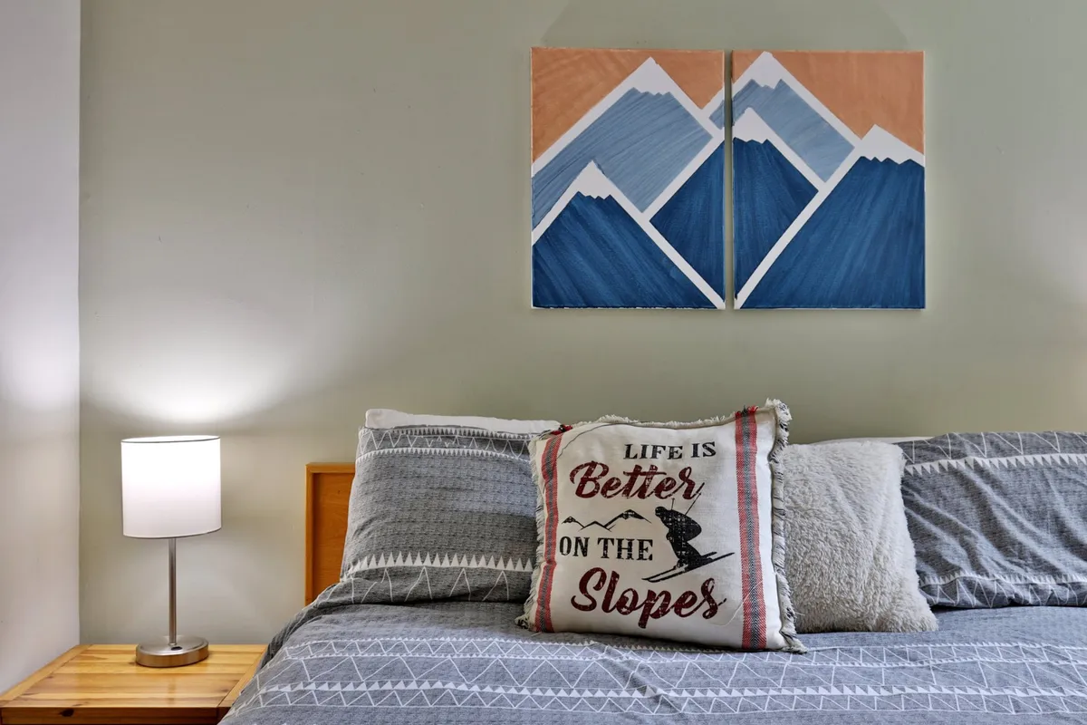 A serene bedroom featuring a king-size bed adorned with plush decorative pillows, mountain-themed artwork above the headboard, and a lamp on the bedside table. The bedding appears cozy and inviting, hinting at a comfortable night's rest.