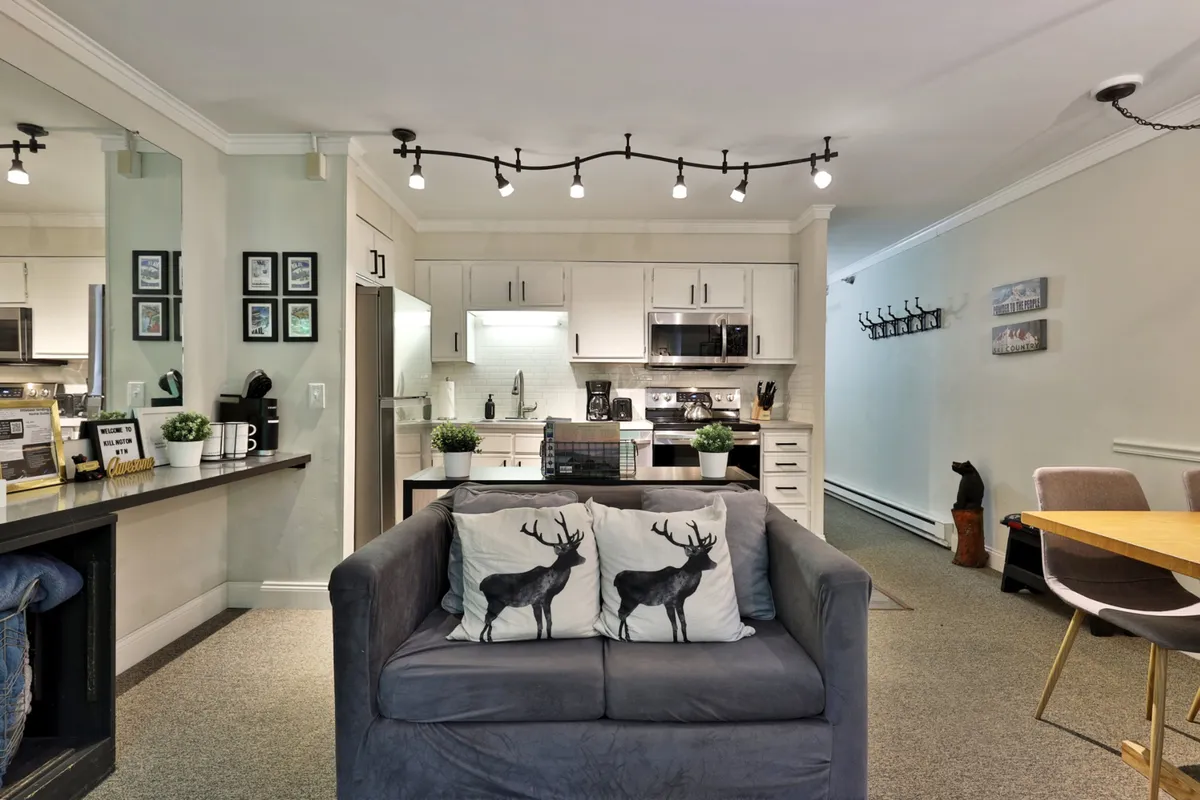 A cozy, open-concept living space combines sightlines of the kitchen, dining table, and a sofa with playful moose-patterned pillows. The layout shows how the kitchen flows seamlessly into the dining and relaxation areas, fostering easy conversation and post-dinner relaxation.