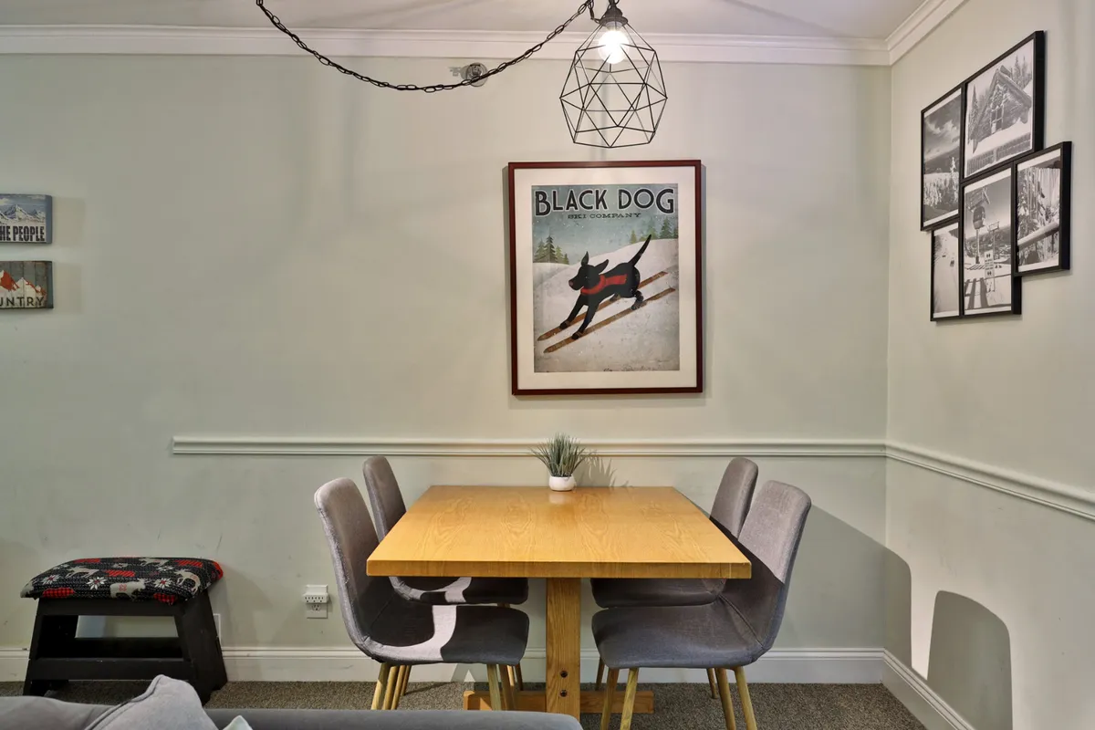 This image captures a warm and welcoming dining area, featuring a substantial table surrounded by chairs. Overhead, a chandelier adds an inviting glow, while artwork on the wall personalizes the space. The table is central, signaling a place for shared meals, games, and lively conversation.