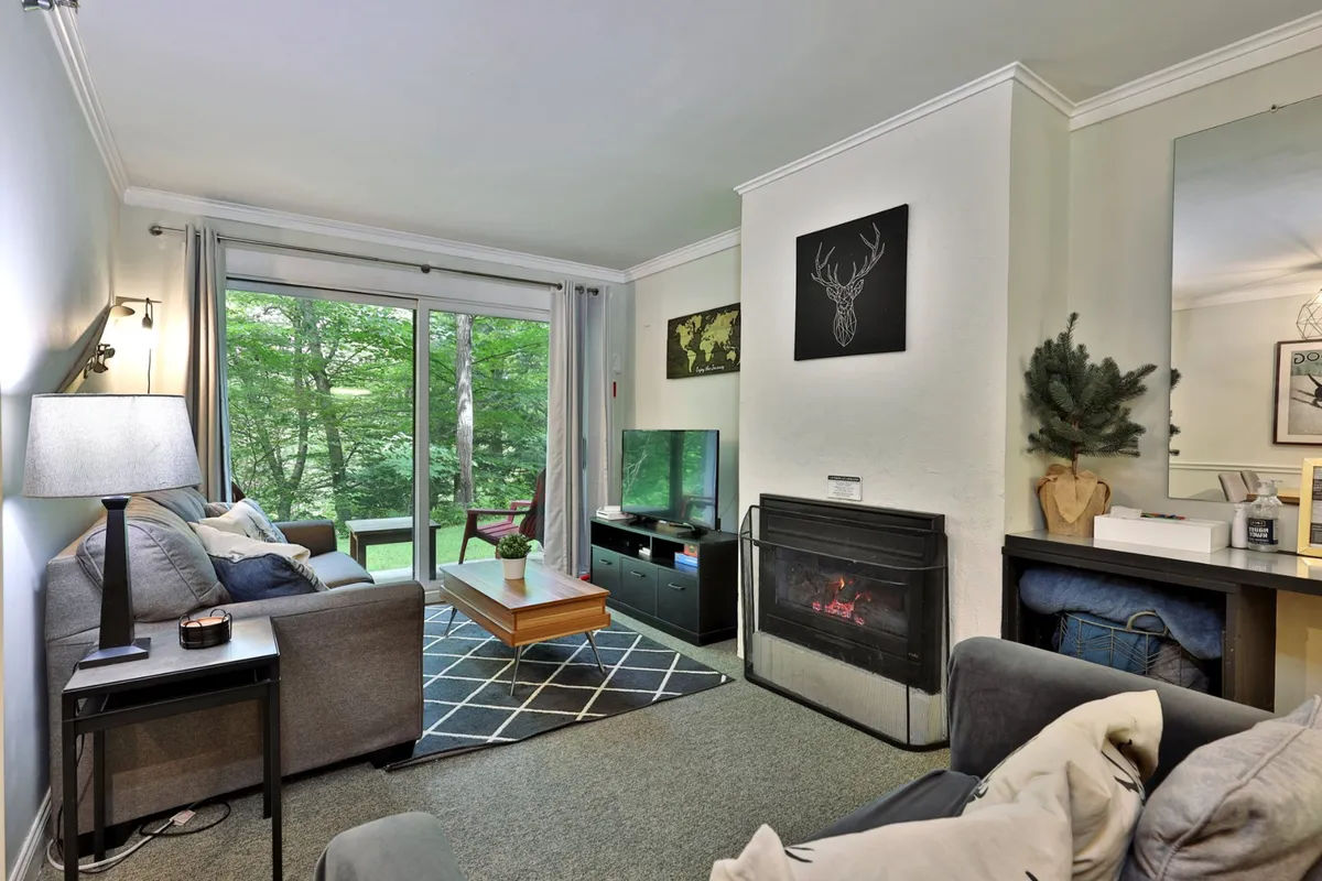 The image showcases a cozy and inviting living area in the Whiffletree E2 condo. A plush sofa sits beside a warm, crackling fireplace, with a stylish coffee table in front. Large windows flood the space with natural light, offering views of the Vermont landscape. The overall ambiance is both relaxing and welcoming, perfect for unwinding after a day on the slopes.