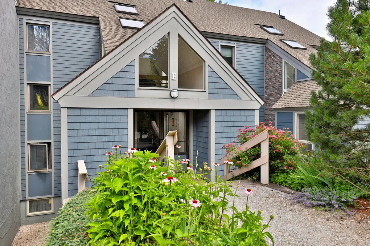 The image displays the entrance of Little Bear Landing with large windows, vibrant flower beds, and inviting wooden steps. The exterior exudes a welcoming atmosphere, suggesting a place families are eager to return to after a day of adventure. The garden and porch area hint at seasonal enjoyment, from summer blooms to fall foliage.