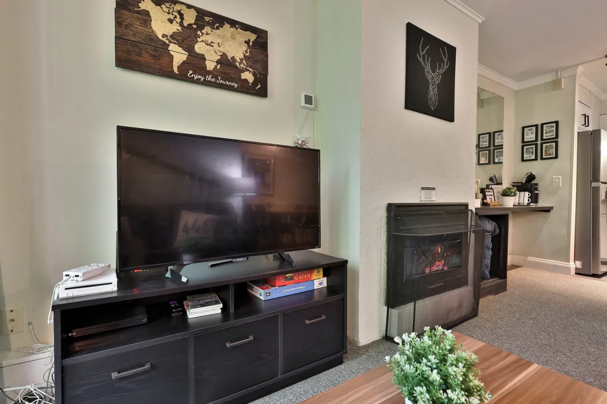 A comfortable living room scene featuring a television, wooden world map wall décor, a fireplace with a decorative deer head, and a coffee table with a small plant. The seating area is arranged for group activities like movie nights or board games.